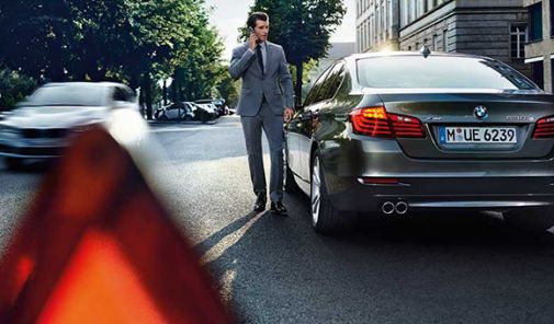 rotes Warndreieck im Vordergrund, ein Mann telefoniert mit seinem Smartphone und steht dabei neben seinem BMW 5er im, im Hintergrund