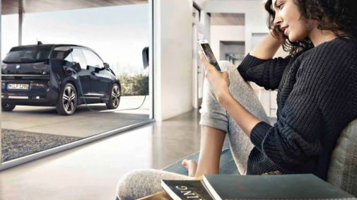 Frau sitzt mit Smartphone bequem im Wohnzimmer auf Ihrer Couch während draußen Ihr schwarzer BMW i3 geladen wird