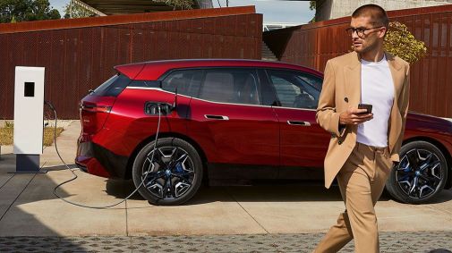 Mann mit beigem Anzug mit einem Smartphone in der rechten Hand vor seinem roten elektrischen BMW Modell